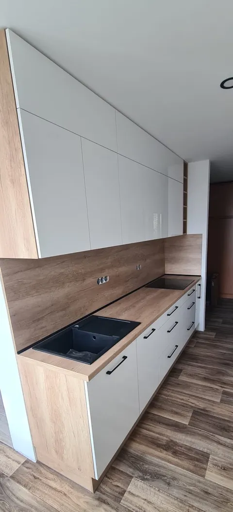 Kuchyňská linka podél stěny, bílá lesklá dvířka, dubová pracovní deska a zástěna, černý dřez, černá varná deska a černá madla